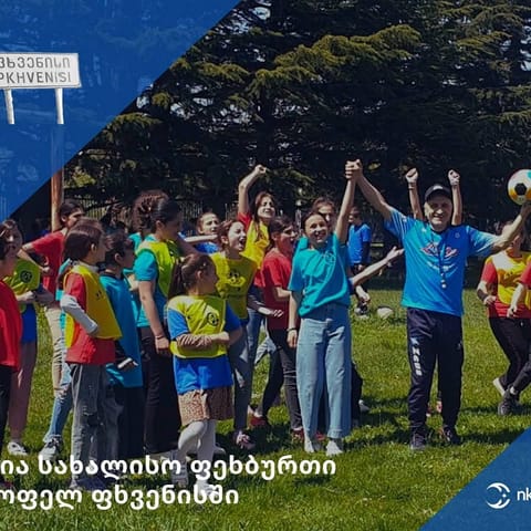 ფერად მაისურებსა და ყვითელ ჟილეტებში გამოწყობილი ბავშვები და ბურთით ხელში მამაკაცი ხელებაწეულები ზეიმობენ მინდორზე; კუთხეში სოფლის დაფა „ფხვენისი“, ქვემოთ წარწერა „ღია სახალისო ფეხბურთი სოფელ ფხვენისში“ და nkfund.-ის ლოგო